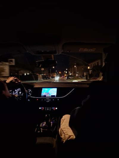 A nighttime photograph taken from inside a moving vehicle depicts a street scene in Hainstadt, Germany. The driver's right hand is visible on the steering wheel, which displays an illuminated dashboard showing a speed of 45 km/h. A central infotainment screen shows a navigation map with a blue route, indicating an estimated arrival time of 5 minutes and a distance of 3.9 km. Below the screen, illuminated climate controls display "23 auto" and media buttons. An automatic gear shifter is visible on the center console. To the right, the silhouette of a front-seat passenger is discernible. Outside the vehicle, the road is illuminated by streetlights, revealing buildings on both sides. Other vehicles with illuminated headlights and taillights are present on the road. A circular blue traffic sign with a white arrow indicating a mandatory right turn is visible on the left side of the road. Motion blur on the left side of the image suggests the vehicle is in motion. The passenger-side sun visor is present, featuring an "AIRBAG" warning label.