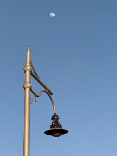 This image, captured in Safaga, Egypt, presents a minimalist composition featuring an ornate street lamp fixture against a clear blue sky, with the moon visible in the upper left corner.

The dominant subject is a metallic lamp arm, possibly brass or a gold-painted metal, which extends horizontally from the right side of the frame. The arm features intricate curves, decorative elements, and cylindrical sections, suggesting a classic or antique design. The metal surface appears textured or slightly weathered, catching the light. At the far end of the arm, a dark, downward-facing lamp head with a distinct shade is visible.

To the upper left, the moon is prominently displayed against the vivid blue sky. It is a bright, gibbous moon, not full, with visible craters and surface details, indicating it's a daytime or twilight sighting. The sky itself is a uniform, cloudless blue, providing a stark and expansive background for the two distinct elements.

The composition utilizes significant negative space, drawing attention to the stark contrast between the man-made fixture and the natural celestial body. There are no people or other objects in the frame, emphasizing the quiet, serene atmosphere. The presence of the moon in a bright blue sky suggests the photograph was taken during daylight hours, perhaps early morning or late afternoon. No visible text is present in the image.