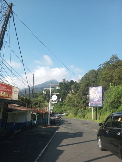 The image shows a road in a rural area, with a mountain in the background. On the left side of the road, there are small shops with rusty roofs. A billboard promoting "SEDAAPNYA FAVORIT SEMUA" and "WARUNG IBU TUTI" is visible. Further down the road, there's a decorative street lamp with the word "AL ALIM" on it. On the right side of the road, there's a billboard advertising "Old Italian" and "Rest Park", noting "4.7 Km". A black car is partially visible on the right side of the road. The environment appears to be a roadway in a mountainous area, surrounded by trees. The sky is blue with scattered clouds. There is no specific indication of the location as Phouviang-Gnai, Laos.