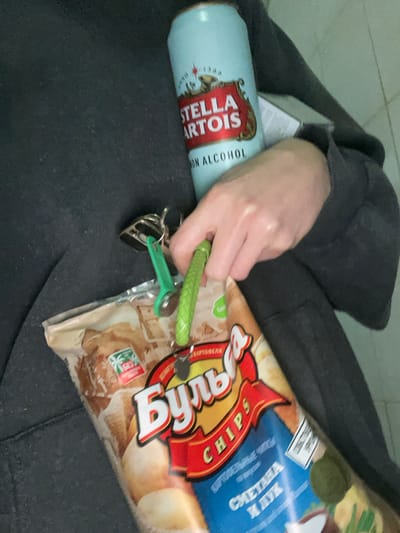 The image shows a person wearing a dark grey hoodie holding a can of Stella Artois Non Alcohol beer and a bag of "Бульба" potato chips in their hand. They are holding a set of keys, which are attached to a green strap. The background is a tiled wall. The coordinates indicate a location in Москва, Russia.