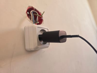 A black power adapter is plugged into a white electrical outlet mounted on a lightly textured, off-white wall. Above and behind the outlet, exposed electrical wires in red, blue, and green are visible within a red circular junction box that is not fully enclosed by the wall. The outlet itself appears to be part of an unfinished or partially installed electrical system. The lighting is soft, casting faint shadows. The image appears to be a close-up shot, focusing on the electrical components and the wall. There are no people present in the image. The scene is set in a domestic or construction environment. The specific location in the city of Avtury, Russia cannot be determined from the visual information provided.