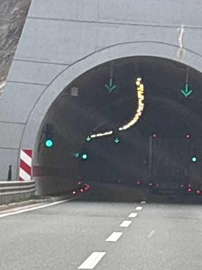 A concrete arch tunnel entrance is observed from a driver's perspective on an asphalt road in Donje Mrke, Montenegro. The tunnel exterior is gray, constructed from segmented blocks. To the left of the entrance, a red and white striped hazard marker stands beside a metal guardrail. The interior of the tunnel is dark, illuminated by various traffic guidance lights. Three green downward-pointing arrows are visible overhead, along with two distinct green circular lights on the lower left and right walls. A prominent curved line of small, illuminated yellow lights arches from the lower left towards the upper right within the tunnel's ceiling. Below these, multiple small red circular lights are visible along the bottom edge of the tunnel's interior. The asphalt road in the foreground features white dashed lane markings leading directly into the tunnel. No people or specific vehicles are discernible.