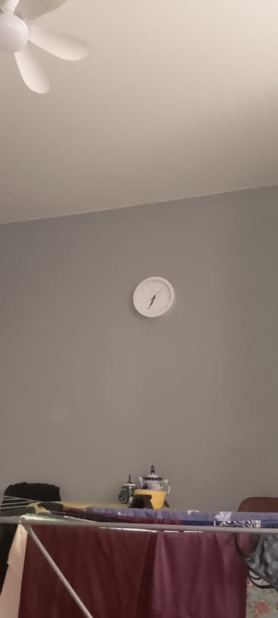 A white, circular analog clock with black hands is mounted on a plain grey wall. The minute hand points to approximately 12, and the hour hand points to approximately 1. Below the clock, a clothes drying rack is visible, draped with various colored fabrics, including a dark red one in the foreground. On a shelf above the drying rack, several objects are present: a small white and blue teapot, a green container, and a yellow cup. In the upper left corner of the image, a portion of a white ceiling fan with three blades is visible. The overall scene appears to be an indoor domestic setting.