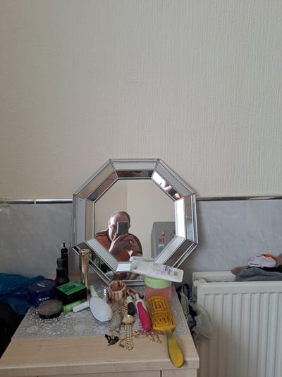 The image depicts a vanity dresser or tabletop with various personal items, predominantly makeup and toiletries, arranged in a slightly cluttered manner. In the center, an octagonal mirror with a beveled, mirrored frame reflects a middle-aged man taking the photo with his smartphone. The man is wearing an orange and black horizontally striped t-shirt and glasses. Behind the mirror, a portion of a small refrigerator with magnets on its door is visible.
On the surface of the dresser, to the left, there is a blue plastic bag, a blue Nivea cream tub, and a smaller round tin. To the right of these are several bottles and containers, including a dark bottle with a narrow neck, a green box, and a gold-colored spray bottle. Further to the right, a white electric toothbrush with a pink head and a yellow hairbrush are prominent. A gold-colored wristwatch with a link band and several strands of black beads are also scattered on the surface. A roll of toilet paper is partially visible on the far right, next to a white radiator covered with folded clothes. The background consists of a textured, light-colored wallpaper above a tiled section of the wall.
The scene appears to be an indoor domestic setting, likely a bedroom or bathroom, specifically located within a residence in the town of Hyndburn, United Kingdom. The overall impression is one of everyday use and personal grooming, with a self-portrait taken in a mirror.