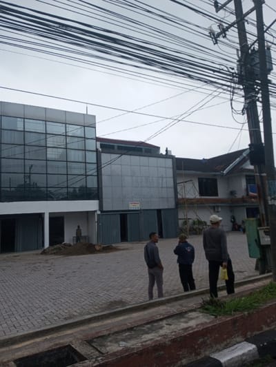A commercial building with large mirrored windows is the main focus. Three men stand on the paved driveway in front of the building. In the background is another building in need of repair. Overhead, a tangle of power lines stretches across the sky, connected to a utility pole on the right. The sky is overcast, suggesting a cloudy day. A sidewalk and curb run along the bottom of the frame.