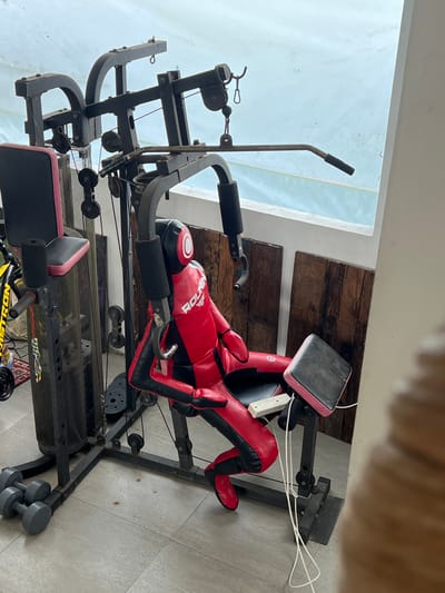 The image shows an indoor setting, likely a room in a house or building located in Singakerta, Indonesia. The room features a home gym setup. The primary subject is a weight machine. Other objects present include a pair of dumbbells. The environment is a well-lit interior space with a tiled floor, and partial view to what appears to be an outdoor water feature or swimming pool. There are no people visible in the image. The actions and interactions are unobservable due to the inanimate nature of the scene and the absence of living beings.