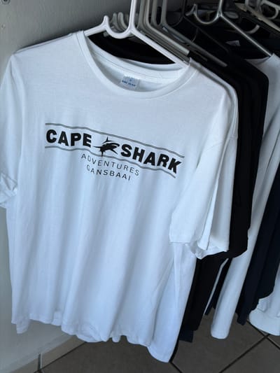 The photograph displays a stack of neatly folded and hung T-shirts, primarily white with some black ones interspersed, suspended on a mix of white plastic and silver metal hangers. The shirts are presented from a slightly elevated, angled perspective, with the foreground dominated by a white T-shirt featuring a prominent black logo. The logo reads "CAPE SHARK ADVENTURES" in bold, capitalized letters, with a stylized shark silhouette positioned between "CAPE" and "SHARK". Below this, smaller text indicates "GANSBAAI". A small white label visible on the collar of one of the white T-shirts shows the brand "VIC BAY" and the size "S".
The T-shirts appear to be merchandise on display, likely in a retail setting or a gift shop. The background suggests an indoor or covered environment, possibly a wall and a tiled floor. There are no people visible, and no specific time of day or weather conditions can be discerned. The "GANSBAAI" text on the T-shirt directly correlates with the provided location context of Overstrand Local Municipality, South Africa, indicating these are local souvenirs or branded apparel related to activities in that area, likely shark diving or viewing adventures.