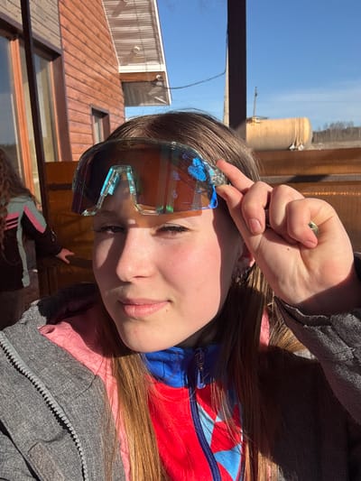 A young person with light skin and light brown hair is posing for a selfie. They are wearing futuristic-looking sunglasses with blue accents. Their left hand is raised near their forehead, partially obscuring the top of the glasses. They are wearing a gray jacket over a pink shirt and a red and blue zip-up top.
The environment outside features architectural structures, including a building with a corrugated metal roof. Visible in the background is a cylindrical tank, likely part of an industrial complex. The sky is clear and blue. The specific location within Gory, Russia, cannot be determined based solely on this image.