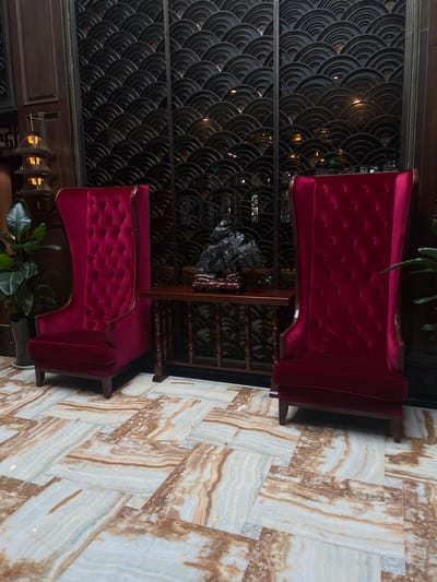 The image displays an interior setting, likely a hotel lobby or lounge, located in Tongxiang, China. Two identical high-back armchairs, upholstered in deep red tufted velvet with visible wooden frames, are centrally positioned. A dark wooden console table sits between the chairs, supporting a black, irregularly shaped ornamental rock displayed on a carved wooden base. Behind the furniture, a dark decorative screen or wall panel extends, featuring a repetitive scalloped pattern. To the left of the left armchair, a tall potted plant with broad green leaves is visible, adjacent to a dark wooden column housing a multi-tiered, pagoda-style light fixture. The floor is covered in large, polished stone tiles exhibiting white, cream, and brown veining, arranged in a rectilinear pattern. No individuals, actions, or interactions are present within the frame.