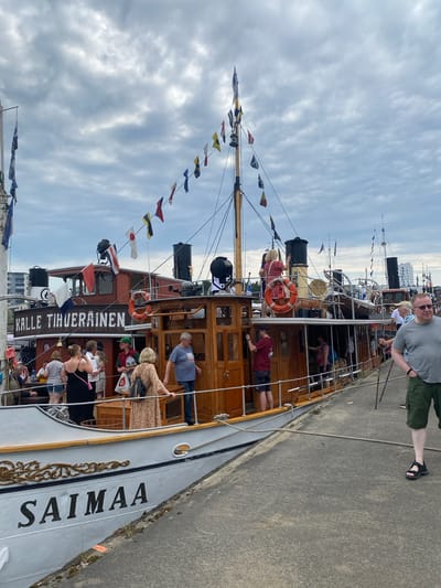The photograph depicts the historic steamship "Saimaa" docked at the Joensuu harbor in Finland. The ship, painted white with ornate gold detailing and black lettering that reads "SAIMAA," is the primary subject. Several people are visible on the ship's deck and the adjacent pier. On the ship, some individuals are standing and interacting, with one woman in a patterned dress leaning on a railing. The ship itself is a multi-decked vessel with a prominent wooden cabin structure, adorned with various nautical flags strung across its masts. To the right of the ship, a man in a grey t-shirt and green shorts stands on the concrete pier, holding two walking sticks. The background shows the Joensuu cityscape with buildings and a cloudy sky overhead. The scene suggests a public event or a day of maritime activity at the harbor.