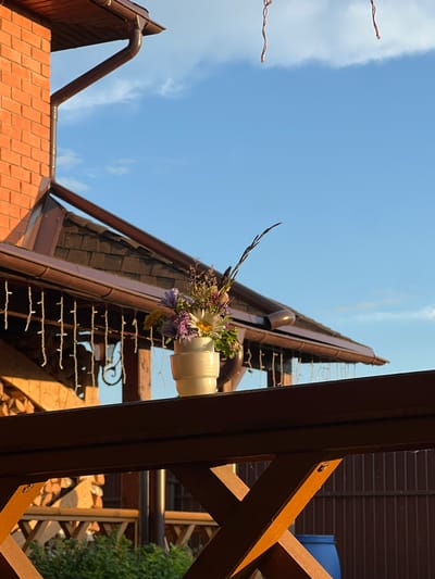 An outdoor scene in Bol'shaya Shishovka, Russia, features a bouquet of wildflowers in a white vase placed on a wooden railing. Behind the railing, a brick building and a wooden roof with dark shingles are visible. The sky is clear blue with a few white clouds. The lighting suggests it is late afternoon or early evening, with warm sunlight illuminating the scene.