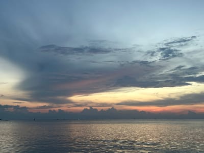 The image depicts a seascape at sunset in Baan Wang Ta Kien, Thailand. The sky is filled with layers of clouds, transitioning from shades of blue at the top to yellow, orange, and grey hues closer to the horizon. A band of dark, cumulonimbus clouds sits near the water's edge. The sea reflects the colors of the sky, with ripples and waves creating a shimmering effect. Small boats are visible on the horizon.
