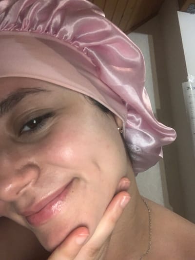 A close-up captures a person with fair skin and dark hair wearing a pink satin bonnet. The person is turned sideways, with their face tilted towards the camera. Their right hand is gently touching their face near the mouth. The focus highlights soft features and a slightly oily sheen on the skin. A delicate silver chain is visible around the neck. The background is a blurred mix of light and dark tones.