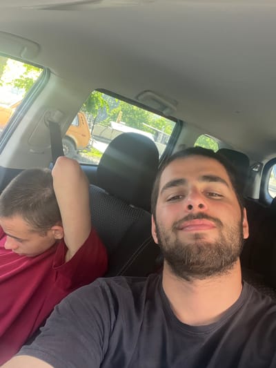 Two young men are seated inside a vehicle, possibly a SUV, in Stepantsminda, Georgia. The man in the foreground, on the right, is taking a selfie. He has short dark hair, a beard, and is wearing a dark t-shirt. The man in the background, on the left, is looking away from the camera and has his arm raised behind his head. He has short brown hair and is wearing a maroon t-shirt. The interior of the car appears to be a standard vehicle interior with fabric seats and a seatbelt visible. Outside the car, through the windows, lush green trees and an orange vehicle can be seen, suggesting an outdoor, possibly rural, setting within the city.