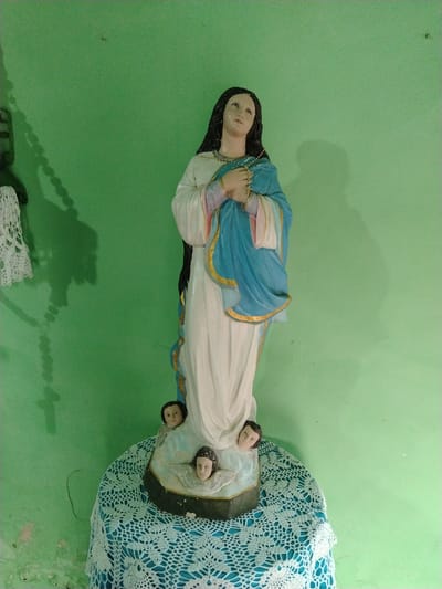 A statue of the Virgin Mary stands on a table draped with a white and blue crocheted tablecloth. The statue is primarily white with a blue and gold robe, and has dark hair. At the base of the statue are three cherubic figures. Behind the statue is a light green wall. To the left of the statue, a portion of a green cross and white crocheted item are visible on the wall. The location is Los Leoncitos, Venezuela.