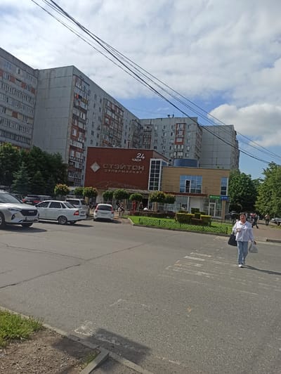 A woman is crossing a crosswalk in front of a supermarket in Dariali, Georgia. She is wearing a white shirt and blue jeans and carrying two shopping bags. The supermarket is named "Стоитон Супермаркет" and is open 24 hours. There are several cars parked in front of the supermarket and a large apartment building in the background. The sky is blue with some clouds.