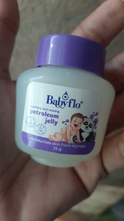 A close-up shot shows a small, white plastic bottle of Babyflo petroleum jelly held by a person's hands. The bottle has a purple cap and a label with the brand name "Babyflo" in blue, accompanied by a droplet logo. Below the brand name, text reads "soothing and relaxing petroleum jelly." The label also features an illustration of a smiling baby sitting next to a panda. At the bottom of the label, it states "Moisturizes skin from dryness 25 g." The background is blurred, showing a textured surface that appears to be a wooden table. The image is well-lit, highlighting the product and the hands holding it. The location in Kawit, Philippines is not visually discernible from the image itself.
