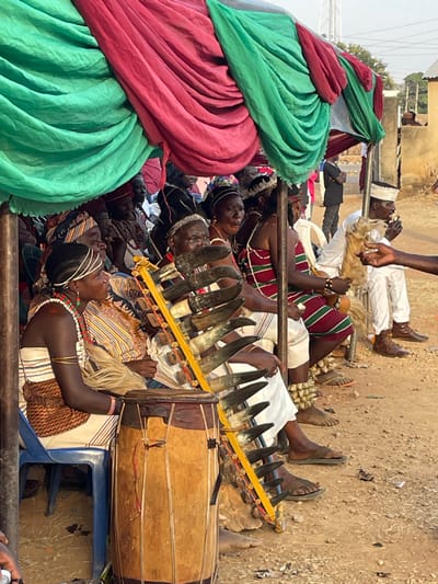 The image captures a vibrant outdoor cultural gathering, presented in a portrait orientation where the subjects appear to be lying on their sides, suggesting the original photo was taken with the camera rotated or the subjects are actually reclining.
In the foreground, taking up the left side of the frame, are large swathes of fabric in bright green and reddish-pink, meticulously draped and folded. These textiles form a colorful, textured backdrop or part of a temporary structure.
Beneath and extending to the right of these fabrics is a diverse group of people, primarily adults with dark complexions, engaged in an event. Many are reclining or lying down on what appears to be a raised platform or ground covering. They are adorned in various traditional or ceremonial garments. Several individuals wear striped fabrics in patterns of red, green, black, and white. Some have elaborate head coverings or intricate hairstyles, and many are decorated with beads and jewelry around their necks and arms.
A prominent musical ensemble is central to the scene. A unique instrument composed of numerous animal horns (at least 10-12) is arranged horizontally on a yellow wooden frame, resembling a type of horn xylophone or panpipe. Next to it, a large, cylindrical wooden drum, crafted from lighter and darker wood sections, rests on a blue plastic chair, ready for use.
Among the people, one individual in the lower right section of the image (when viewed upright) is distinctly visible, reclining with a striking light-colored, possibly braided or wig-like, hairstyle and multiple strands of colorful beads. Further back, another person in a white robe and cap, holding what appears to be a light-colored animal fur or tassel, is visible. The bare feet of several individuals are seen, some wearing sandals, others boots.
The setting appears to be outdoors on dry, light brown ground or dirt. In the background, hints of other structures, possibly buildings, and distant vegetation are visible under what seems to be bright daylight. The gathering, traditional attire, and musical instruments suggest a ceremonial performance, festival, or cultural celebration is taking place. The relaxed, reclining postures might indicate a period of rest during a long event or a customary way of observing the proceedings.