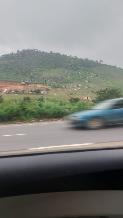 The image shows a landscape scene with a blurred blue car in the foreground, indicating motion. The car is on a road with a white line marking its lane. In the background, there is a green, hilly landscape with scattered trees and rocks. There are some buildings visible on the hillside, appearing to be under construction or with a rustic appearance. The sky is overcast. The scene appears to be in a rural or semi-rural area, possibly outside of a city like Dam Camp, Nigeria, given the presence of natural terrain and less developed structures.