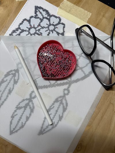 The image depicts a close-up view of a diamond painting project in progress. A white pencil lies diagonally across a translucent canvas printed with a design of leaves and a floral element. To the right of the pencil, a pink heart-shaped tray holds a variety of small, faceted beads in shades of gray and rose gold. In the upper right quadrant of the image, a pair of black-rimmed glasses rests on the canvas. The project is laid out on a wooden surface, with a strip of beige tape securing a portion of the canvas. The lighting suggests an indoor setting, possibly a crafting area. The image does not contain information about the city of Biržai, Lithuania.