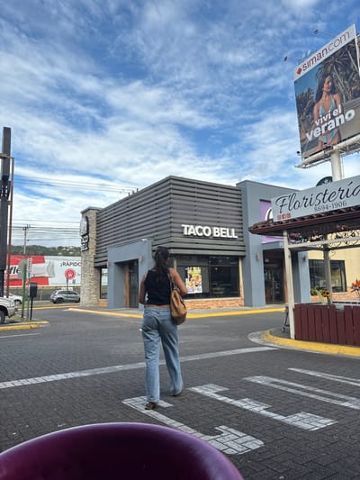 The image shows a woman in blue jeans and a black tank top walking across a crosswalk in front of a Taco Bell restaurant in Pozos, Costa Rica. The restaurant is located at latitude 9.951 and longitude -84.194. There is a large billboard to the right of the restaurant that advertises a clothing store called “Siman.com.” The billboard is showing a woman wearing a bikini and the words “vivi el verano” (live the summer). There is a small, wooden kiosk to the right of the Taco Bell. It is a florist shop with the sign “Floristeria” written in large letters. The sky is blue with white clouds. There are power lines running above the buildings. The woman is walking towards the camera. There are other people in the restaurant, but they are not visible in the image. The crosswalk is marked with white paint. The parking lot is empty except for a car in the background.