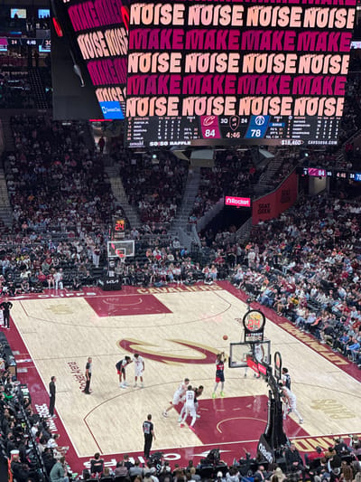 This image depicts a basketball game at Rocket Mortgage FieldHouse in Cleveland, Ohio, U.S.A. The Cleveland Cavaliers are playing a game, with the score being 84 to 78 in the third quarter with 4:46 left on the clock. The arena is filled with spectators, and the scoreboard displays the game information and advertisements. There are multiple players and referees on the court. The main advertising message displayed is "MAKE NOISE".