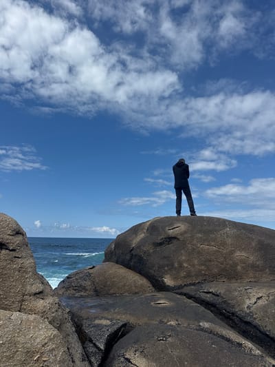 The image shows a person standing on a large, rocky outcrop overlooking the ocean. The person is facing away from the camera, their back to the viewer, wearing a black jacket and dark pants. They are standing on a large, flat, gray rock with an uneven surface, which appears to be part of a larger rock formation. The sky above is a clear blue with white, fluffy clouds scattered across the horizon. The ocean below is a deep blue, with white-capped waves breaking on the shore.  The location is a rocky outcrop, located in the city of Muxia, Spain, at the coordinates 43.113 latitude and -9.221 longitude.