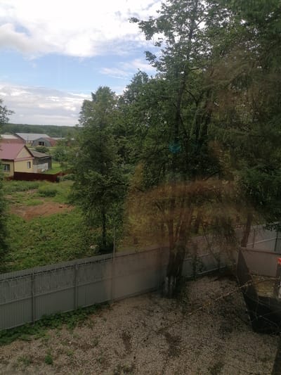 The image depicts an outdoor scene with a rural or suburban setting, likely in the town of Komsomolsky, Russia. The foreground features a gravel ground and a corrugated metal fence topped with barbed wire. Beyond the fence, there is dense green vegetation and several large trees with lush foliage. In the mid-ground, a yellow house with a red roof and a smaller gray outbuilding are visible, surrounded by a red fence and overgrown greenery. In the background, a line of trees forms a horizon, with a few more buildings with reddish roofs and gray structures appearing further back. The sky is partly cloudy with patches of blue. The overall impression is one of a residential area on the outskirts of a settlement, with elements of both nature and human habitation. There are no people or animals visible in the image, and no discernible actions or interactions are occurring.
