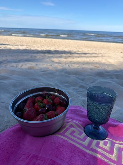 An outdoor shot shows a beach scene with a blue goblet and a bowl of strawberries and cherries placed on a pink towel with a gold Greek key pattern. The beach is made of sand, and the ocean is visible in the background. The sky is blue. The image appears to have been taken during the day, likely in the afternoon, given the lighting. The mood is relaxed and leisurely. The location is Zīvartiņš, Latvia, suggesting a seaside destination. There is no visible text in the image.