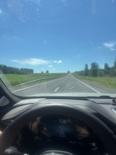 This media file shows a view from inside a car traveling on a highway. The perspective is from the driver's seat, with the steering wheel and dashboard visible in the foreground. The road stretches ahead, bordered by green fields and trees, under a bright blue sky with scattered clouds. The image appears to be taken during the daytime, with clear weather.
The dashboard displays several pieces of information. The dominant numbers show a speed of 120 km/h and a time of 13:51. Other visible text includes "Колеса" (Wheels), "Автопилот" (Autopilot), "Ada D", and a distance of "21330km". There are also some Chinese characters on the screen, "年代金曲(live)", which translate to "Golden Oldies (live)". The reflection on the dashboard suggests there is another person or passengers in the car. The overall mood is one of travel and movement, likely on a road trip. The location context provided is Zeledeevo, Russia.