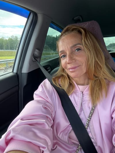 The image shows a woman in a pink satin bomber jacket with a silver chain detail, sitting in the driver's seat of a car. She has blonde hair and is wearing makeup, with a slight smile. A black seatbelt is visible across her chest. The setting appears to be a road trip, with a highway and trees visible through the car windows. The sky is cloudy, suggesting overcast weather. The image is taken from a selfie perspective, with the woman looking at the camera. No visible text is present in the image.