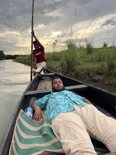 Two men are in a boat on a waterway. In the foreground, a man with a beard and wearing a blue, white, and green patterned short-sleeved shirt and light-colored pants is lying down on a striped mat in the boat. He is looking towards the camera. Behind him, standing in the boat and holding a long wooden pole, is a younger man wearing a red t-shirt with the word "Jaffna" printed in white and light-colored pants with a black stripe. He is looking away from the camera, towards the left side of the frame. The boat is on a river or canal with green vegetation and tall grasses lining the banks. The sky above is cloudy with patches of sunlight breaking through. The scene appears to be in a rural area, potentially near Sylhet, Bangladesh, given the natural landscape and type of boat.