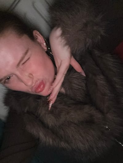 A person with pale skin is lying down against a fuzzy, brown fur coat. The person is positioned at an angle, their head near the bottom left. They have elongated, pointed nails and several rings on their fingers. The person's right hand is raised near their face, and their lips are slightly pursed. Multiple earrings are visible on the left ear. The background is dark and somewhat blurry.