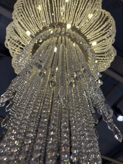 The image depicts a crystal chandelier viewed from below. The chandelier features a circular, flower-like arrangement of lights and crystal strands. Crystal strands cascade downwards, sparkling and reflecting light. The background is dark, suggesting the chandelier is in a dimly lit room. The location is unspecified.