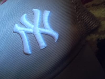 The image shows a close-up view of the embroidered logo on a baseball cap. The logo is white and appears to be the iconic "NY" logo of the New York Yankees. The cap is a light tan color and has a mesh panel on the side, typical of many baseball caps. The lighting is somewhat dim, and the background is out of focus, appearing to be a textured surface, possibly fabric. There is no discernible information about the location in Ezbet Michel Hamawi, Egypt, as the image solely focuses on the cap.
