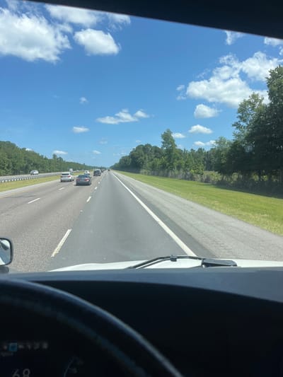 The image is taken from the interior of a vehicle, likely a truck, as evidenced by the steering wheel and dashboard in the foreground and a side mirror on the left. The vehicle is on a highway, with multiple lanes of traffic visible. Other vehicles, including cars and SUVs, are driving on the highway. The environment is a sunny day with a clear blue sky and scattered white clouds. Lush green trees and grass line the sides of the highway, suggesting a rural or suburban setting. The road appears to be in good condition, with clear lane markings. The time of day appears to be daytime, based on the bright sunlight and shadows. There is no visible indication of the specific location in the city of Riceboro, United States, in the image itself.