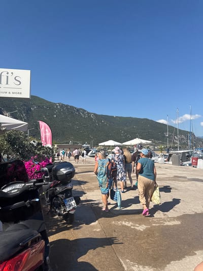 In the Municipal Unit of Kamena Vourla, Greece, a group of people are walking along a paved waterfront area on a sunny day. The scene is characterized by a clear blue sky and a verdant, mountainous backdrop. Several boats are docked in the harbor, with masts and sails visible. In the foreground, a black scooter with a license plate reading "HYE 353" is parked. To the left, a sign with the text "fi's" and "RIUM & MORE" is visible, along with flowering pink bushes. The individuals are casually dressed, some carrying shopping bags and beach towels, suggesting they are on vacation or returning from an outing. The overall atmosphere is relaxed and typical of a seaside town.