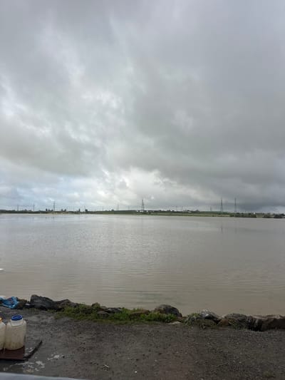 The scene, located in Oumazza, Morocco, depicts an expansive body of murky, brownish-gray water under a uniformly overcast sky. The sky is filled with dense, light gray clouds, showing no visible sunlight. The water surface exhibits subtle ripples, extending from the immediate foreground to the distant horizon. Along the far shoreline, a low, flat landmass is visible, punctuated by numerous tall, metal lattice-work electricity transmission towers, some partially obscured by a hazy atmosphere. Scattered green vegetation or trees are present along this distant bank. In the immediate foreground, a dark, granular ground surface, likely gravel or dirt, runs horizontally across the bottom of the frame. This ground abuts a low embankment constructed of dark, irregular rocks and interspersed with small patches of green grass and weeds. On the far left of the foreground, two light-colored, possibly plastic, jugs with blue caps are visible, resting on a dark, flat surface, alongside a crumpled blue plastic bag. A white, flat object, potentially a table edge, is partially visible at the extreme bottom left corner. No individuals or active interactions are observed in the image.