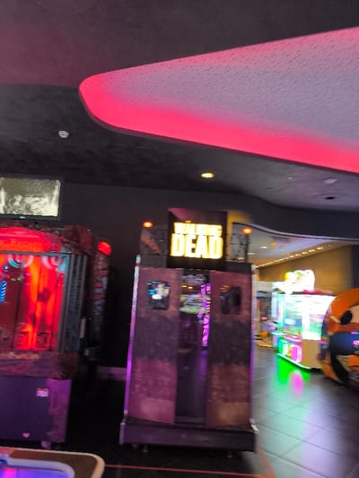 The image captures the interior of an arcade or entertainment center in Agios Dimitrios, Greece, characterized by a dimly lit environment illuminated by vibrant electronic game displays and architectural lighting. The ceiling is dark and textured, featuring a prominent, curved white panel with a continuous strip of bright red light running along its edge, casting a strong red glow across the space.

In the midground, a large arcade game prominently displays the yellow, capitalized text "THE WALKING DEAD" on a marquee, indicating it is likely a game based on the popular zombie-themed franchise. This machine has a rugged, dark appearance with side panels that suggest an immersive gaming experience. To its left, another large arcade machine glows intensely with red and blue lights, though its specific game title is obscured. Above this machine, a smaller monitor displays a grainy, perhaps vintage or distorted, monochrome image.

Further into the background on the right, several other arcade games are visible, emitting a spectrum of neon colors including green, pink, and blue, creating a dynamic light show. Part of a character from the Disney "Cars" franchise is also discernible on one of the machines in the far right. The floor is dark and reflective, contributing to the vibrant, artificial lighting atmosphere. No people are visible, but the setup clearly indicates a venue for gaming activities, likely active during the evening or night.