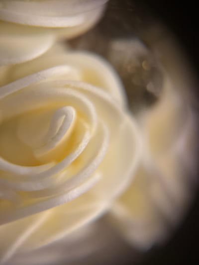 The image is a macro photograph of a white swirl of frosting, resembling a rose petal or a flower. The frosting is creamy and appears to be a light yellow-white color. The texture is smooth with subtle ridges and curves, indicative of being piped. The lighting creates highlights and shadows, emphasizing the three-dimensional form of the frosting. The background is dark and out of focus, with some bokeh effects in the upper right quadrant, suggesting a shallow depth of field. The setting is not identifiable as being in the city of Biržai, Lithuania, as there are no discernible landmarks or context clues. The image focuses solely on the frosting.