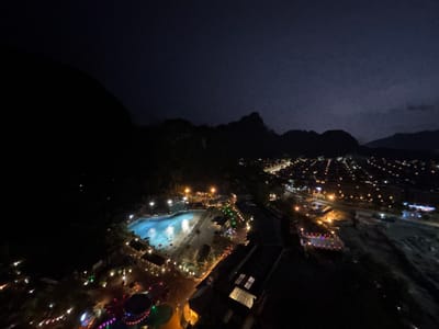 This is an aerial nighttime photograph of a resort complex located in Ipoh, Malaysia. The resort features a large, brightly lit swimming pool with clear blue water, surrounded by loungers and shaded areas. Several buildings with illuminated windows and roofs are visible within the resort grounds, along with pathways lined with decorative lights. A portion of the resort appears to be a water park. Beyond the resort, the lights of a city stretch into the distance, indicating its proximity to an urban area. In the background, dark, silhouetted mountains frame the scene against a deep blue night sky. The image captures the vibrant atmosphere of the resort and the expansive urban landscape of Ipoh at night.