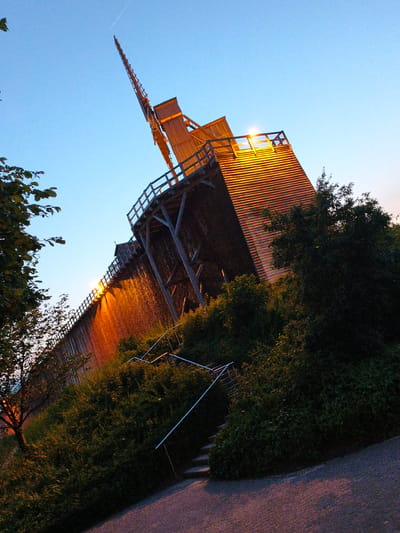 The image shows the Gradierwerk in Bad Rothenfelde, Germany, at dusk. The Gradierwerk is a large, wooden structure designed for salt production. In the image, the structure is illuminated by warm orange lights, highlighting the stacked wooden branches and the water flowing down them, creating a misty effect. The sky above is a clear, soft blue, transitioning into twilight. To the left, a tree with dark green leaves is partially visible, its branches reaching towards the sky. In the foreground and to the right, dense green bushes and trees surround the base of the Gradierwerk. A metal railing and steps are visible, indicating a pathway or access to the structure. The ground is paved with a dark material, possibly asphalt or gravel, with subtle purple and pink hues from the low light. There are no people or animals visible in the image. The perspective is from a low angle, looking up at the imposing structure.