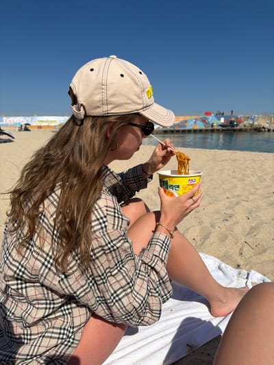 A young woman is sitting on a white towel on a sandy beach, eating noodles from a yellow cup with chopsticks. She is wearing a beige baseball cap, sunglasses, and a plaid shirt dress. The background features a clear blue sky, the ocean, and a colorful structure on the beach. The photograph appears to have been taken at a coastal area with an active beach. The location is possibly in Toseong-myeon, South Korea.