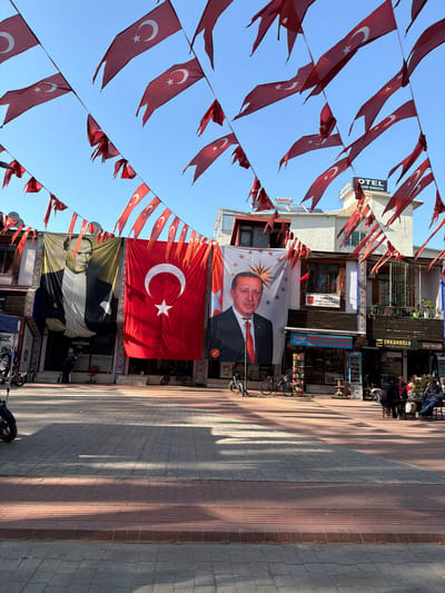 A bustling street in Iznik, Turkey, is captured under a bright, clear blue sky, adorned with national symbols. Numerous small, triangular Turkish flags, featuring the red field with a white star and crescent, are strung across the street, creating an overhead canopy.

Dominating the buildings on the left are large banners. One prominent banner displays a portrait of Turkish President Recep Tayyip Erdoğan against a white background with gold stars. Directly beneath it, an enormous Turkish flag hangs vertically, proudly showcasing its iconic design. Further down, another large banner features a portrait of Mustafa Kemal Atatürk in black and white, partially visible.

The multi-story buildings lining the street house various businesses. Visible signs include "OTEL AND RİMEDYA" and "ERKANOĞLU GAYRİMENKUL." Another sign indicates the "TÜRKİYE MUHARIP GAZİLER DERNEĞİ İZNİK TEMSİLCİLİGİ" (Turkish War Veterans Association Iznik Representation). Other fragmented text such as "Deniz," "Çay Ocağ" (Tea House), "AYSAN," and "ZABITA NOKTASI" (Municipal Police Point) are also visible on storefronts.

On the right side, a paved sidewalk stretches along the street, where a few people are seated at outdoor tables or walking. Bicycles are parked near the shops. Long, distinct shadows are cast across the pavement, suggesting a sunny day, likely late morning or early afternoon. The scene conveys a strong sense of national pride and a lively community atmosphere.