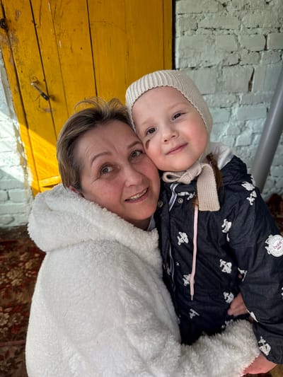 A young child and an adult, presumably the child's grandmother, are embracing in an outdoor setting in Kipchak, Russia. The child is wearing a winter hat and jacket with a distinctive pattern, while the adult is bundled in a white, fleece-like jacket. The background features a building with a yellow wooden section and a white brick portion. A metal pipe runs horizontally across the top of the frame. A section of patterned carpet is visible in the lower right corner of the image.