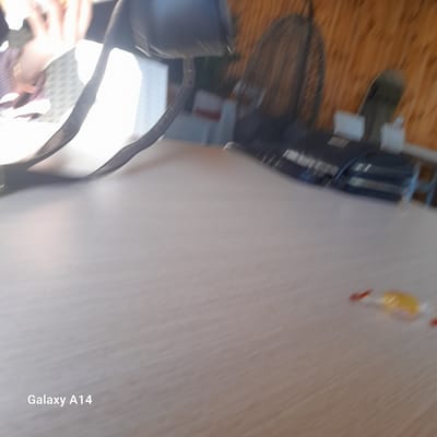 I cannot determine the location in the city of Al-Mawasi or provide precise descriptions of people in this image. The image is blurry, and the orientation is unclear. However, I can see a table, a fan, and what appears to be a purse or bag. There is also a small object that might be a bottle.