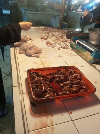 A person is preparing poultry at a market stall. There's a raw chicken on the counter, along with various chicken parts and pieces scattered around. A tray full of chicken livers sits prominently in the foreground, with some blood spilled around the tray. A scale and a metal bowl are also visible on the tiled countertop. The scene appears to be a busy marketplace with other stalls in the background.