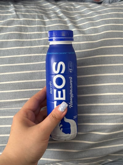 A hand with light skin and a pale blue nail polish with white polka dots is holding a blue plastic bottle with a white label. The bottle has white text that reads "EOS", "ЙОГУРТ", "Натуральный", and "260 г". There are also smaller text elements that appear to be in Russian. The bottle is positioned vertically on a surface with gray and white horizontal stripes. The image is taken from a top-down perspective, showing the hand and bottle against the striped background. The scene appears to be indoors, possibly on a bed or a couch, given the fabric texture. There is no information in the image to suggest this location is in the city of Prikubanskii, Russia.