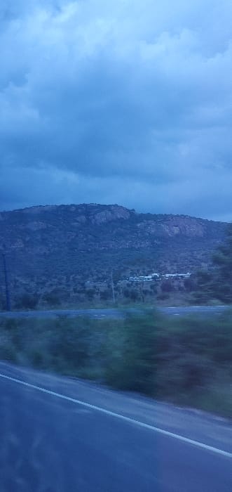 The image displays a rural landscape in Kinanie ward, Kenya, captured during dusk or early night hours under a heavily overcast, dark blue-grey sky. A dark asphalt road, exhibiting motion blur indicative of a moving perspective, occupies the lower left portion of the frame, bordered by a single solid white line on its left edge. Blurred dark green vegetation lines the immediate roadside. The mid-ground consists of dense, dark scrub and trees, transitioning into a range of dark, rugged hills and mountains in the background. These hills are characterized by sparse, dark vegetation and exposed rock formations. A cluster of light-colored structures, appearing as buildings, is visible on the lower slopes of the hills in the right-center. No individuals are depicted in the scene.