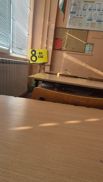 The image depicts a classroom interior in Satovcha, Bulgaria, as indicated by the sign "8-МИ КЛАС" (8th GRADE). Sunlight streams through a window with blinds, casting parallel shadows across the room. The foreground shows the surface of a wooden desk. In the mid-ground, a radiator is visible on the left, and further to the right, a set of tiered desks is arranged. Above the desks, a bulletin board displays educational charts, including one titled "МНОГОСТЕ" (Polyhedrons) with geometric diagrams, and a certificate labeled "ГРАМОТА" (Diploma). A Bulgarian flag is partially visible in the upper right corner. The overall impression is that of a typical Bulgarian classroom.